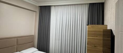 A hotel room with a bed, a bedside table with a kettle, and a large window with curtains.