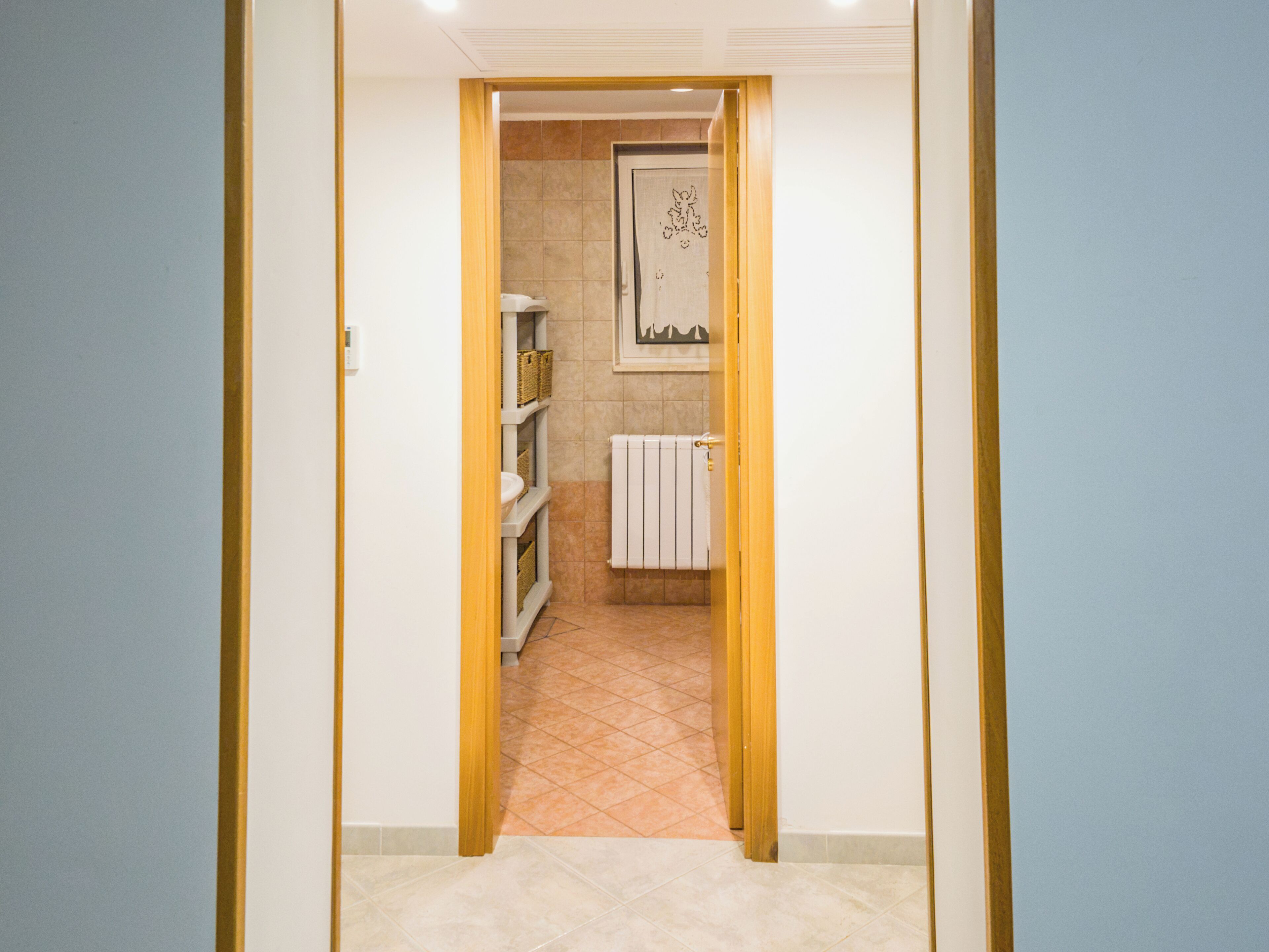 A narrow hallway leading to a bathroom with a radiator, shelves with baskets, and a door with a towel hanging on it.