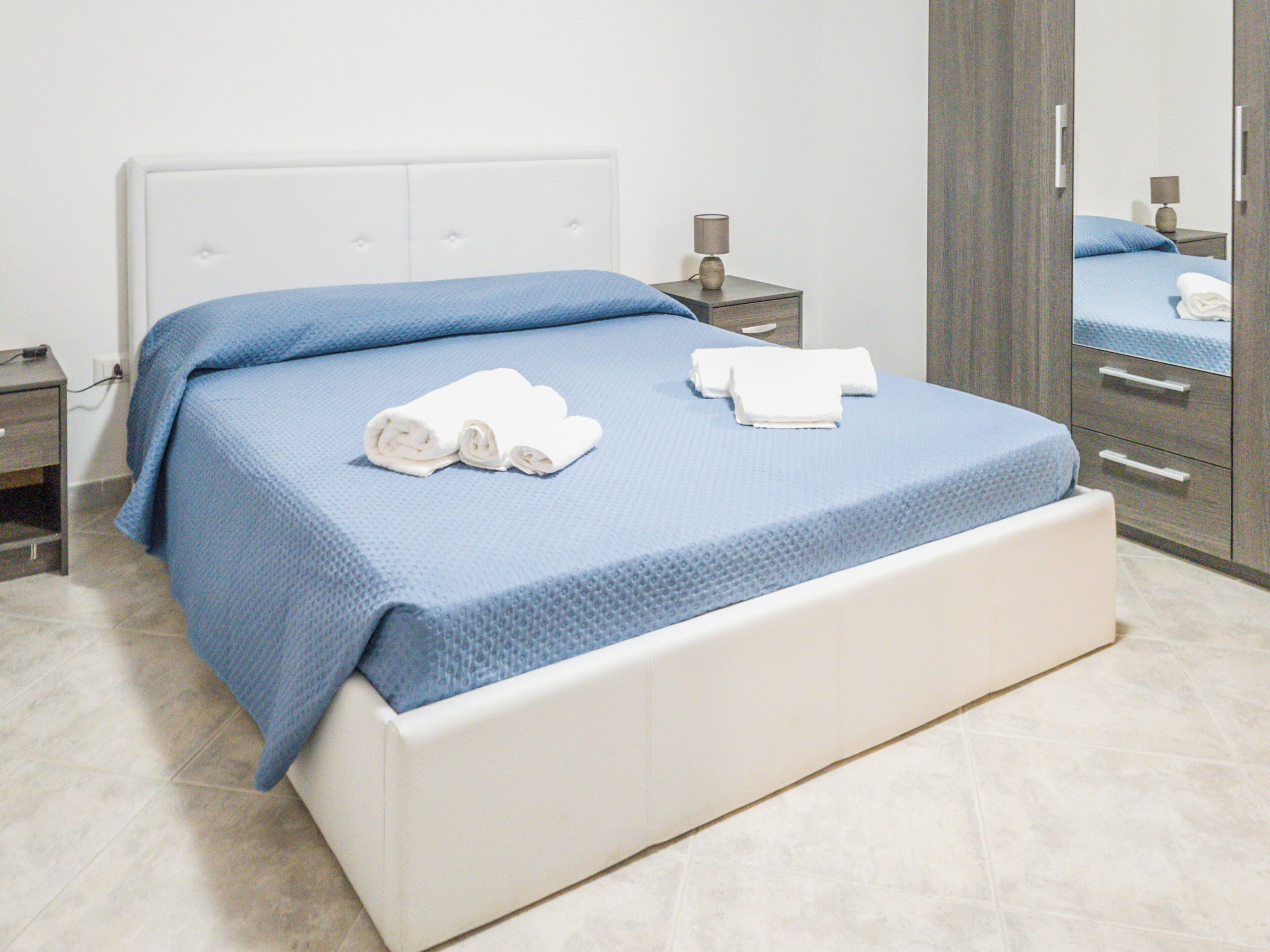 A bed with a blue bedspread and two rolled towels, a bedside table with a lamp, and a wardrobe with a mirror.