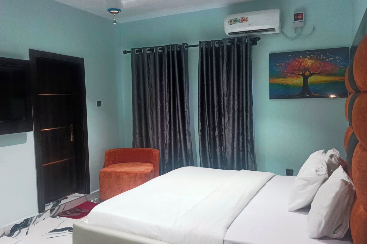 A hotel room with a bed, a chair, a painting, curtains, and an air conditioning unit.