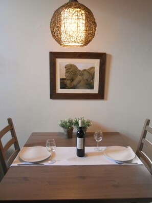A dining table with two plates, a wine bottle, and two glasses, set against a wall with a framed picture and a hanging light fixture. - La Cabane; a comfortable, detached gîte for two with a spacious terrace (Les Bois d'Anjou)