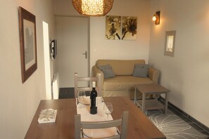Un coin repas avec une table en bois dressée pour deux personnes, une bouteille de vin et un verre d’eau. On aperçoit un canapé beige à l’arrière-plan et un tapis à motifs sur le sol.
