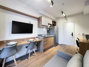 A modern kitchen with a flat-screen TV, wooden cabinets, a stainless steel refrigerator, and a dining area with a sofa and chairs. - My One Estação Japão Liberdade (São Paulo)