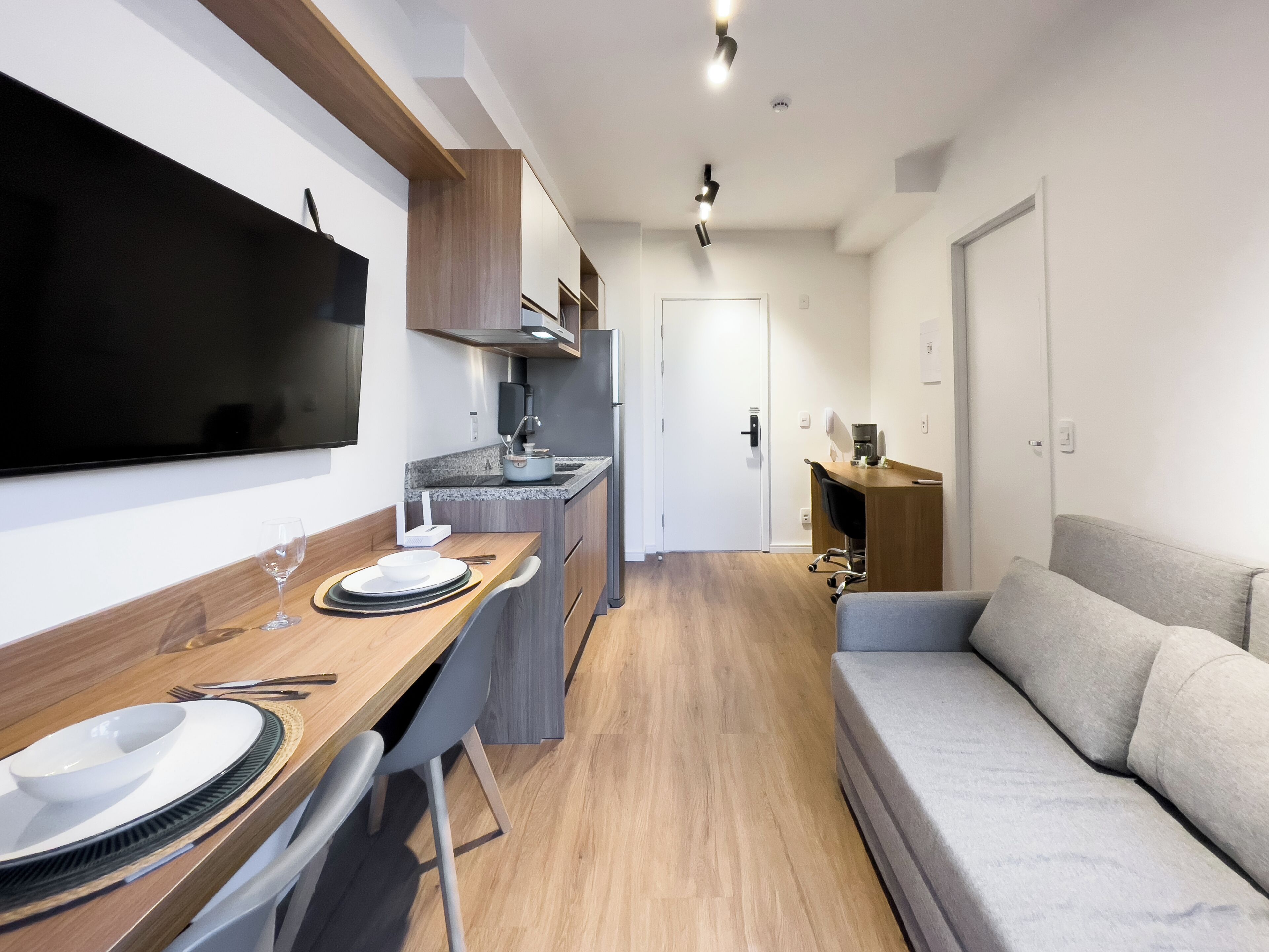 A modern kitchenette with a flat-screen TV, a dining area with a wooden table and chairs, and a grey sofa.
