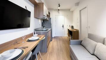 A modern kitchenette with a flat-screen TV, a dining area with a wooden table and chairs, and a grey sofa.
