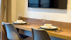 Un comedor con una mesa de madera dispuesta para dos, dos cuencos blancos, dos copas de vino y cubiertos. Un televisor de pantalla plana está montado en la pared sobre la mesa.