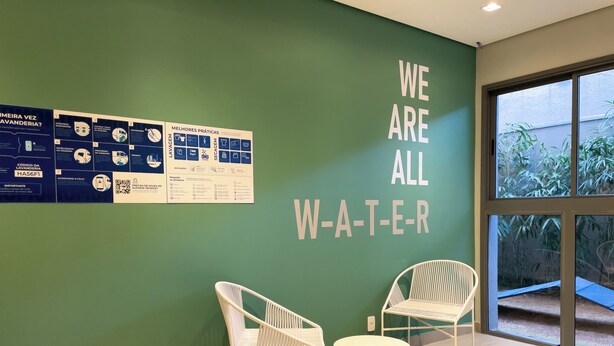 Una habitación con una pared verde, dos sillas blancas, una mesita redonda y un ventanal con vistas a la vegetación.