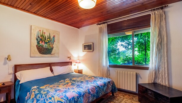 A bedroom with a bed, a nightstand, a lamp, a window with curtains, a radiator, and a painting on the wall. - Villa with Large Garden and Pool (La Cavada)