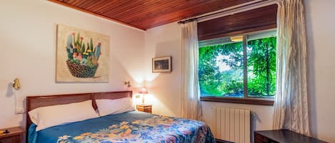 A bedroom with a bed, a nightstand, a lamp, a window with curtains, a radiator, and a painting on the wall.