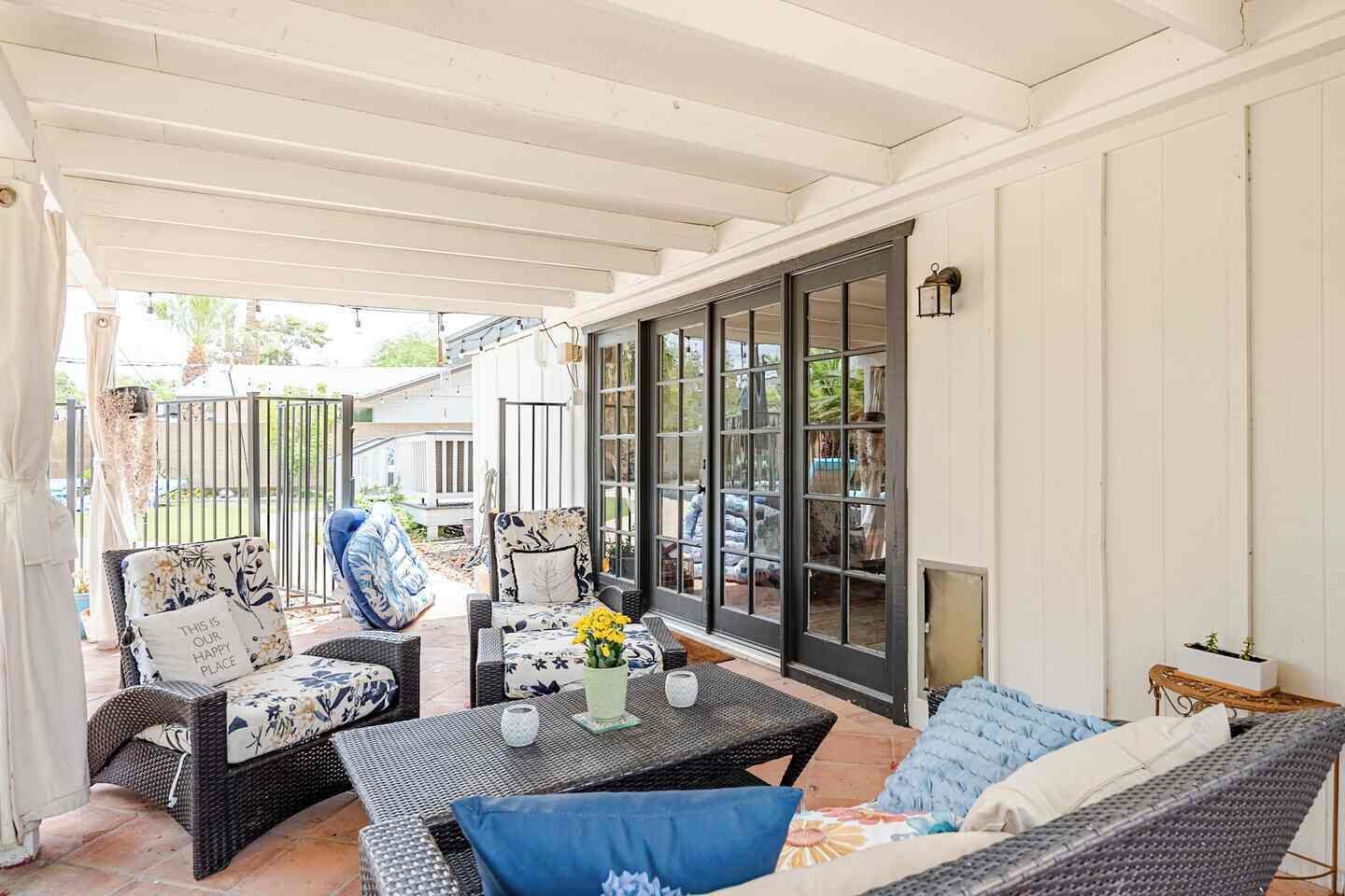 A covered patio area with wicker furniture, a table with a vase of flowers, and a view of the backyard through large sliding glass doors.
