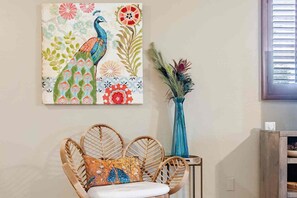 Una habitación con una silla de mimbre, un jarrón azul con plantas secas y un cuadro de un pavo real colorido en la pared.