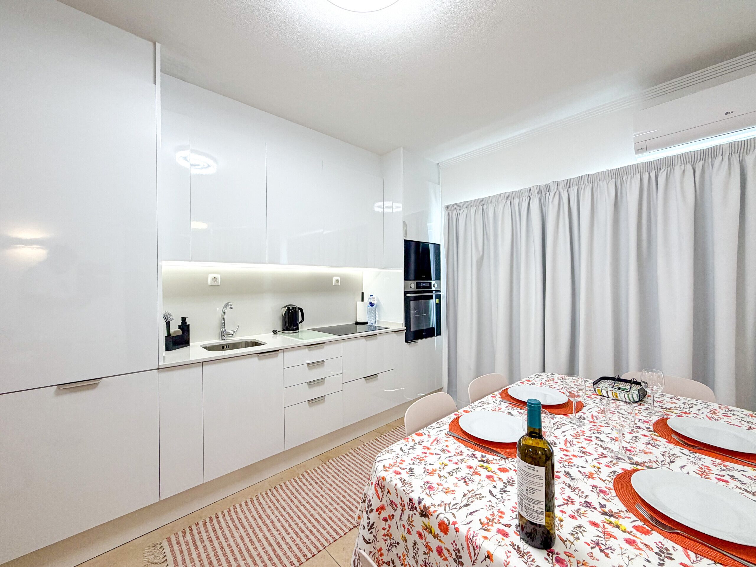 A modern kitchen with white cabinets, a built-in oven, and a dining area with a floral tablecloth and red placemats.