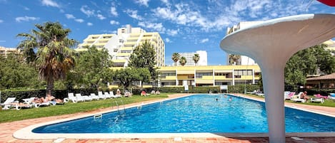Uma piscina com um deck revestido, rodeada por espreguiçadeiras e palmeiras.