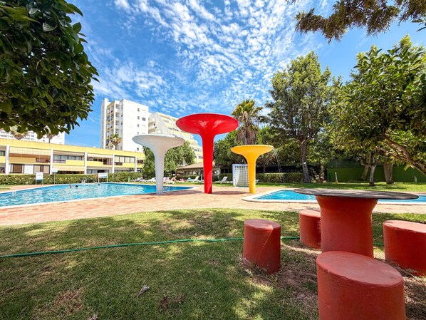 A swimming pool with colorful abstract sculptures, a grassy area, and a building in the background. - Apartment Praia Rocha | Pool | Modern Vogue (Portimão)