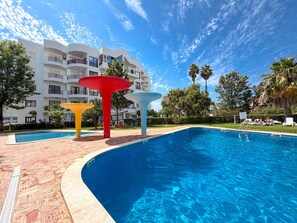 A swimming pool with a modern design, surrounded by a landscaped area with palm trees and a multi-story building in the background. - Apartment Praia Rocha | Pool | Modern Vogue (Portimão)