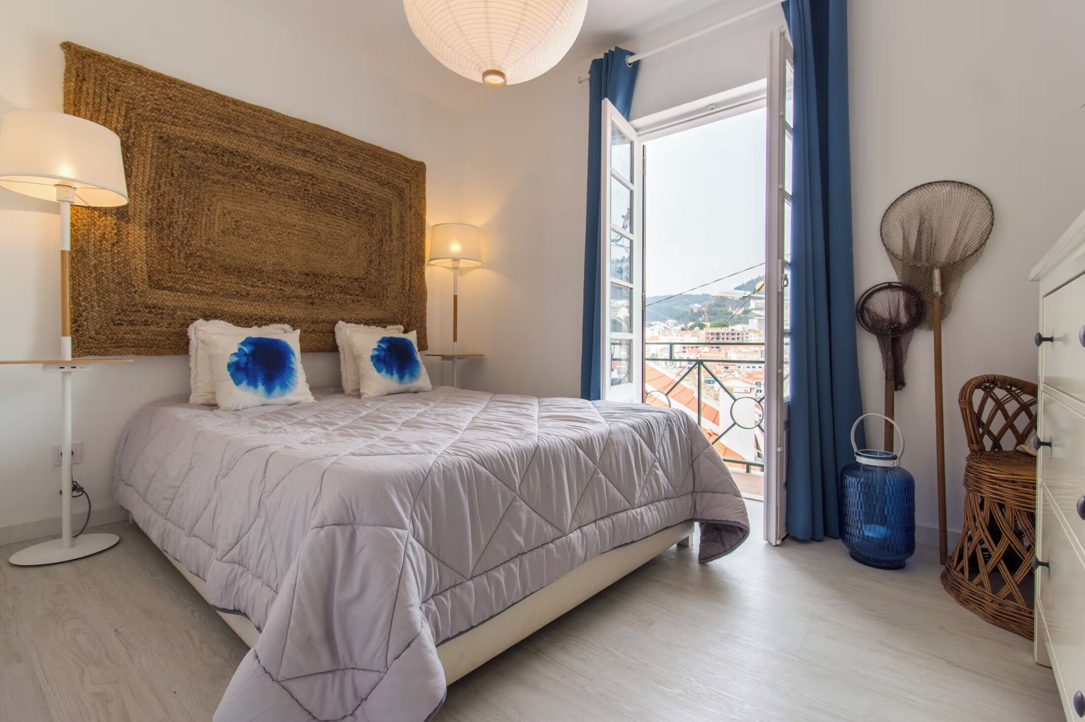 Une chambre à coucher avec un lit, une lampe de chevet, une grande tête de lit en osier tressé, une vue par la fenêtre et un fauteuil en rotin.