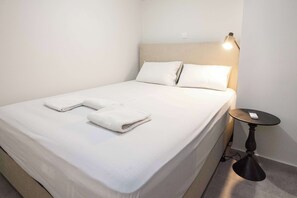 Ein ordentlich bezogenes Bett mit weißen Leinen und zwei gefalteten Handtüchern, ein Nachttisch mit einer Lampe und eine Steckdose an der Wand.