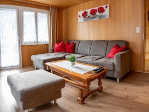 A living room with a grey sectional sofa, red cushions, a wooden coffee table with a vase and remote controls, and a grey ottoman. - Bergfreude Appartement/Fewo 4-Bettwohnung by Interhome (Saas-Grund)