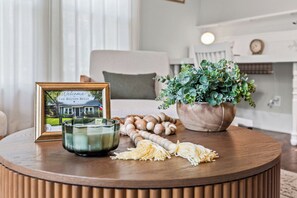 Une pancarte d’accueil encadrée, une bougie et une plante sont posés sur une table en bois dans une pièce où se trouvent un lit et une horloge au mur.