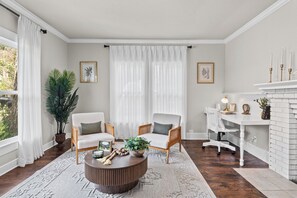 Ein Wohnzimmer mit rundem Couchtisch, zwei Sesseln, einem Beistelltisch, einem Kamin und einem großen Fenster mit Vorhängen.