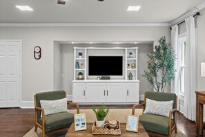 Un saló amb un televisor de pantalla plana, dues butaques tapissades de verd, una taula de centre amb una planta i una biblioteca amb objectes decoratius.