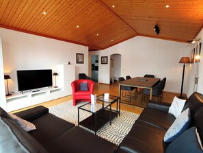 Ein modernes Wohnzimmer mit Fernseher, einem roten Sofa, schwarzen Ledercouchs, einem Esstisch mit Stühlen und einer Küche im Hintergrund.
