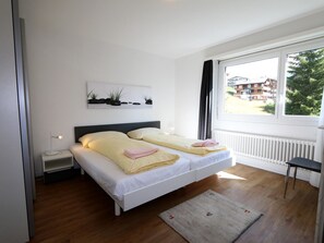 A bedroom with a bed, bedside table, lamp, window, and a view of buildings outside. - Cristal Appartement/Fewo 6-Bettwohnung Nr. 8 by Interhome (Saas-Fee)