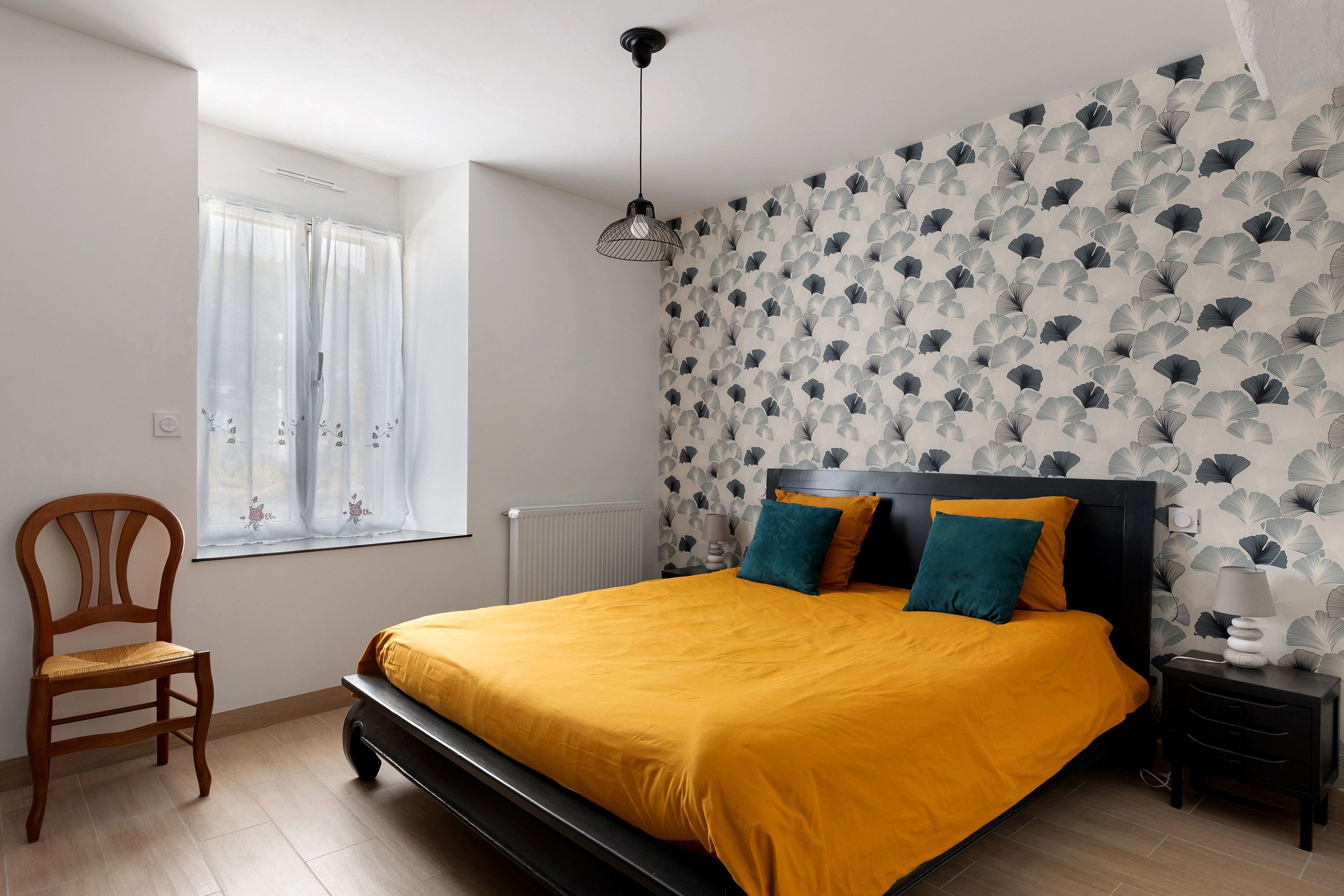Ein Schlafzimmer mit Bett, Sessel, Nachttisch, Lampe und einem Fenster mit Vorhängen.