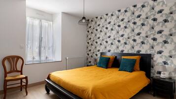 Ein Schlafzimmer mit Bett, Sessel, Nachttisch, Lampe und einem Fenster mit Vorhängen.