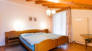 A bedroom with a wooden bed, a small bedside table, a chair, a round table, and a window with curtains. - Haus Evita Appartement/Fewo 4-Bettwohnung Nr. 60 by Interhome (Saas-Grund)
