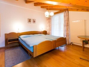 Ein Schlafzimmer mit einem hölzernen Bett, einem kleinen Nachttisch, einem Stuhl, einem runden Tisch und einem Fenster mit Vorhängen.