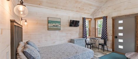 A wooden cabin room with a bed, desk, and chair, a television, and a door.