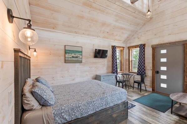 A wooden cabin room with a bed, desk, and chair, a television, and a door.