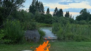 A campfire surrounded by rocks in a grassy area with a path leading to a body of water and trees in the background. - The Summit Suite | Walk to Rangeley Lake • ATV & Sled Access (Rangeley)