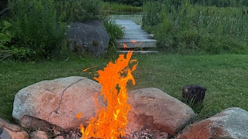 Un foc de campament envoltat de roques en una zona verda amb un camí que condueix a un cos d'aigua i arbres al fons.