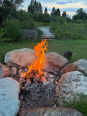 A campfire surrounded by rocks in a grassy area with a path leading to a body of water and trees in the background. - The Summit Suite | Walk to Rangeley Lake • ATV & Sled Access (Rangeley)