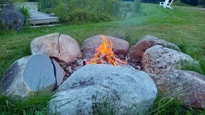 A campfire surrounded by rocks in a grassy field with trees in the background. - The Summit Suite | Walk to Rangeley Lake • ATV & Sled Access (Rangeley)