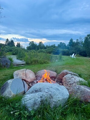 A campfire surrounded by rocks in a grassy field with trees in the background. - The Summit Suite | Walk to Rangeley Lake • ATV & Sled Access (Rangeley)