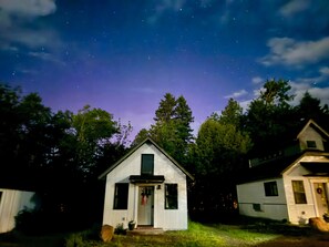 A small, single-story cabin with a front door and a small porch, surrounded by trees and a clear night sky.