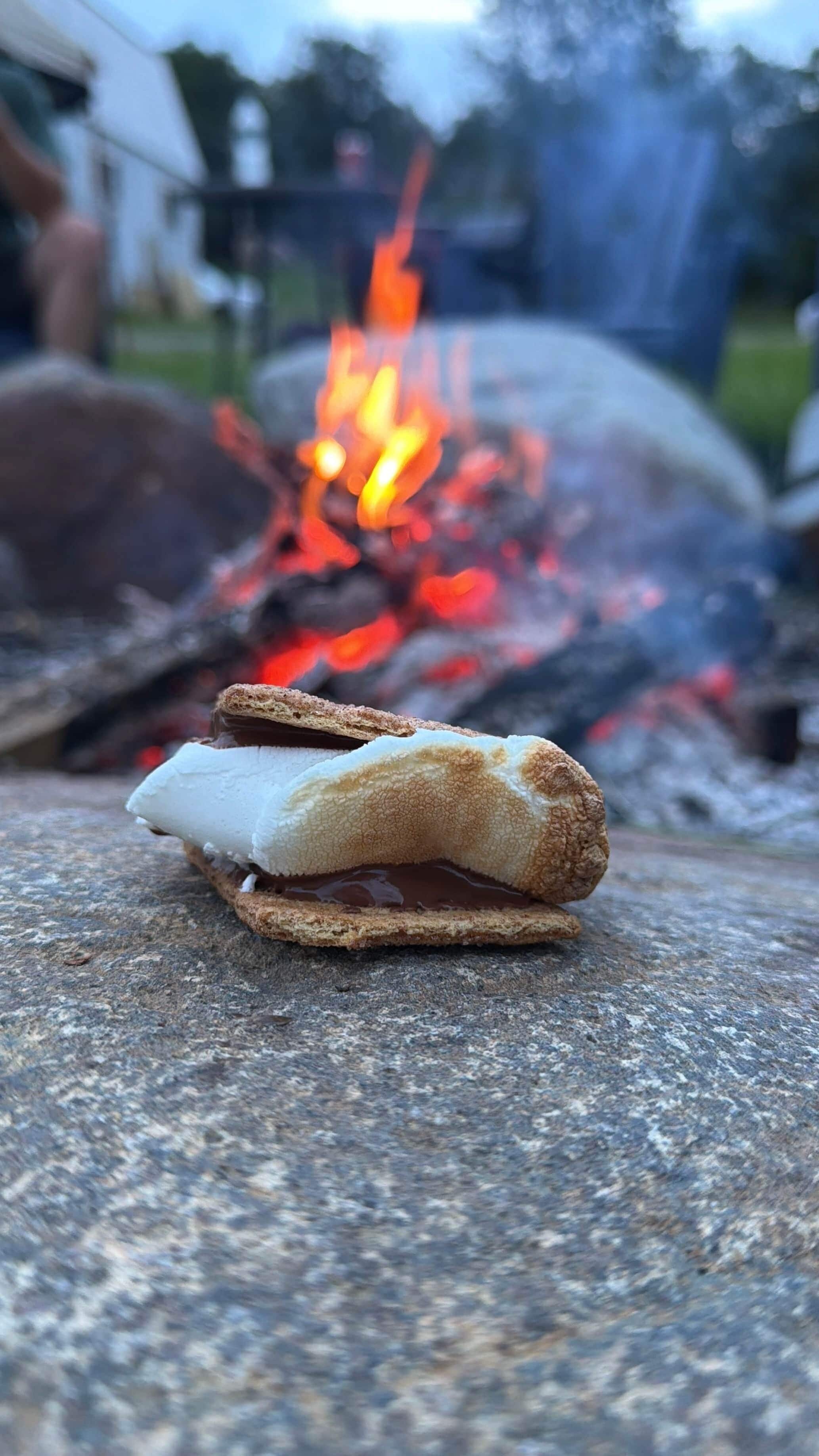 Une guimauve sur un biscuit Graham, posée sur une surface en pierre, avec un feu de camp en arrière-plan.