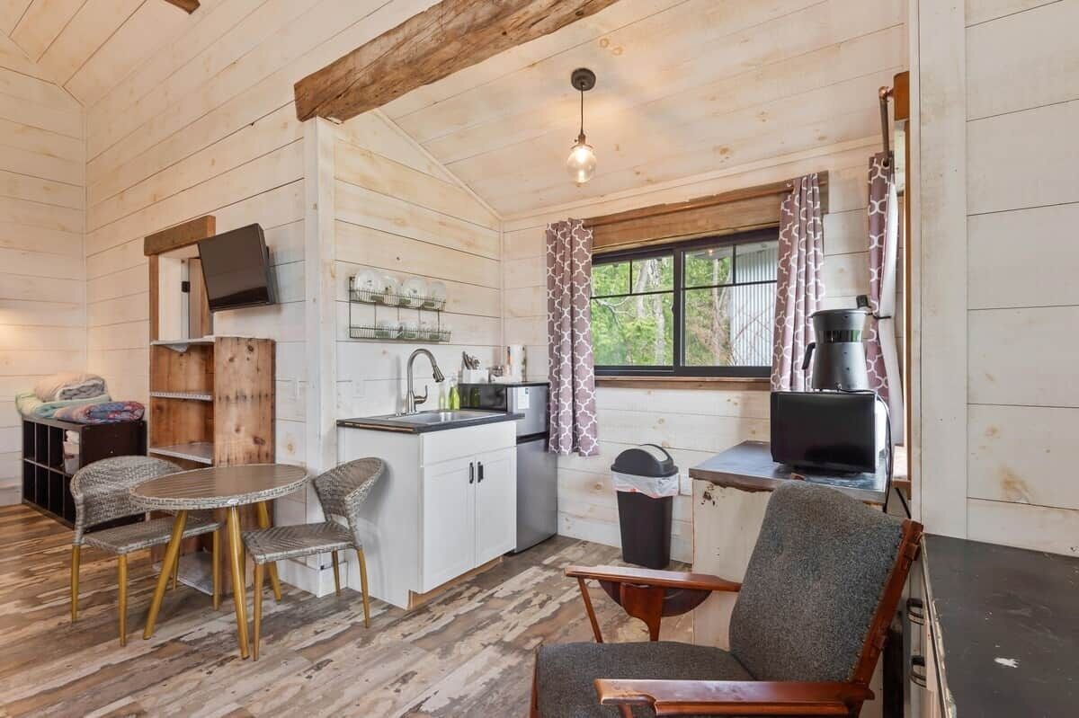 A compact kitchen with a dining area, wooden walls, a window with curtains, and a television mounted on the wall.