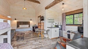 A cabin interior with a bunk bed, a dining area with a table and chairs, a kitchen with a sink and cabinets, and a window with curtains.