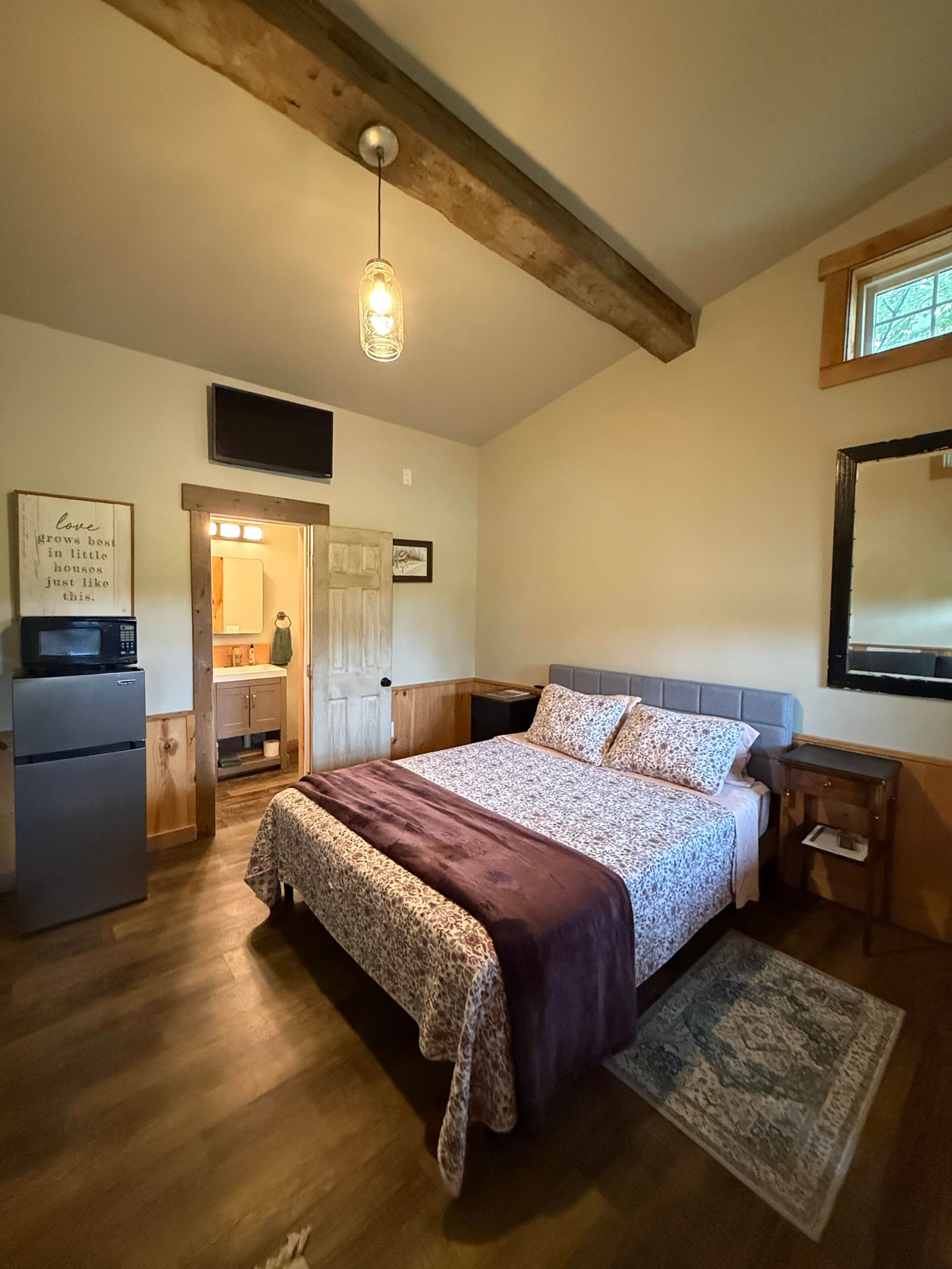 A bedroom with a bed, a nightstand, a wall-mounted TV, a wooden ceiling, and a kitchen area with a refrigerator.