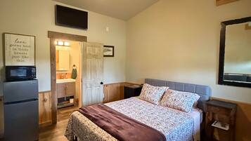 A bedroom with a bed, a nightstand, a wall-mounted TV, a wooden ceiling, and a kitchen area with a refrigerator.