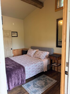 Ein Schlafzimmer mit einem Bett, einem Nachttisch, einem Teppich, einer Tür und einem gerahmten Bild an der Wand.
