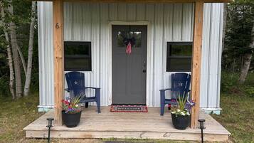 A small, single-story building with a porch, two chairs, and potted plants.