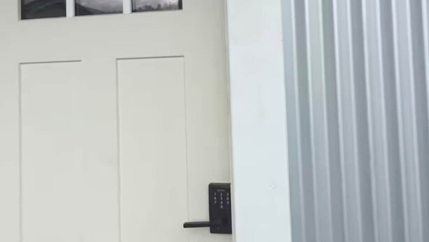 Uma porta branca com fechadura eletrônica, o número '2' na parede e uma janela com vista para um céu nublado.