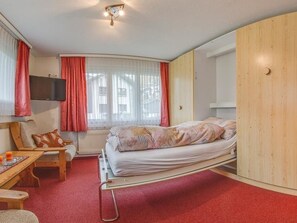 A compact hotel room with a bed, a desk, a chair, a television, and a window with curtains. - Anemone Appartement/Fewo 6-Bettwohnung Erdgeschoss by Interhome (Saas-Fee)