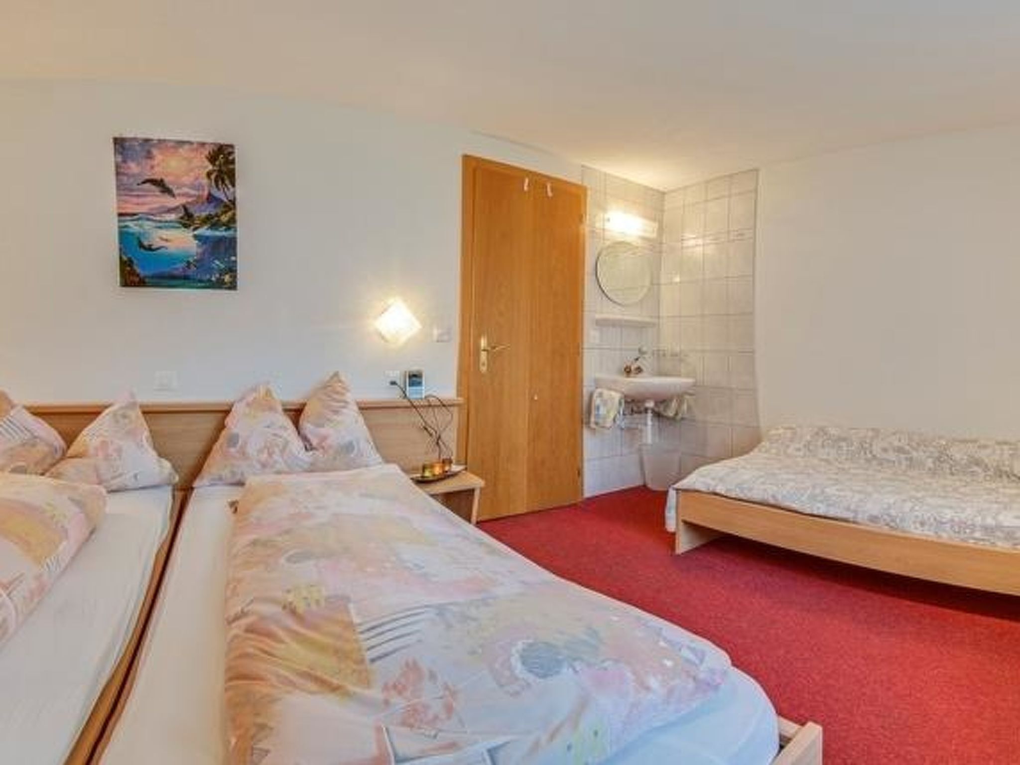 A hotel room with two beds, a wooden door, a bathroom with a sink, and a painting on the wall.