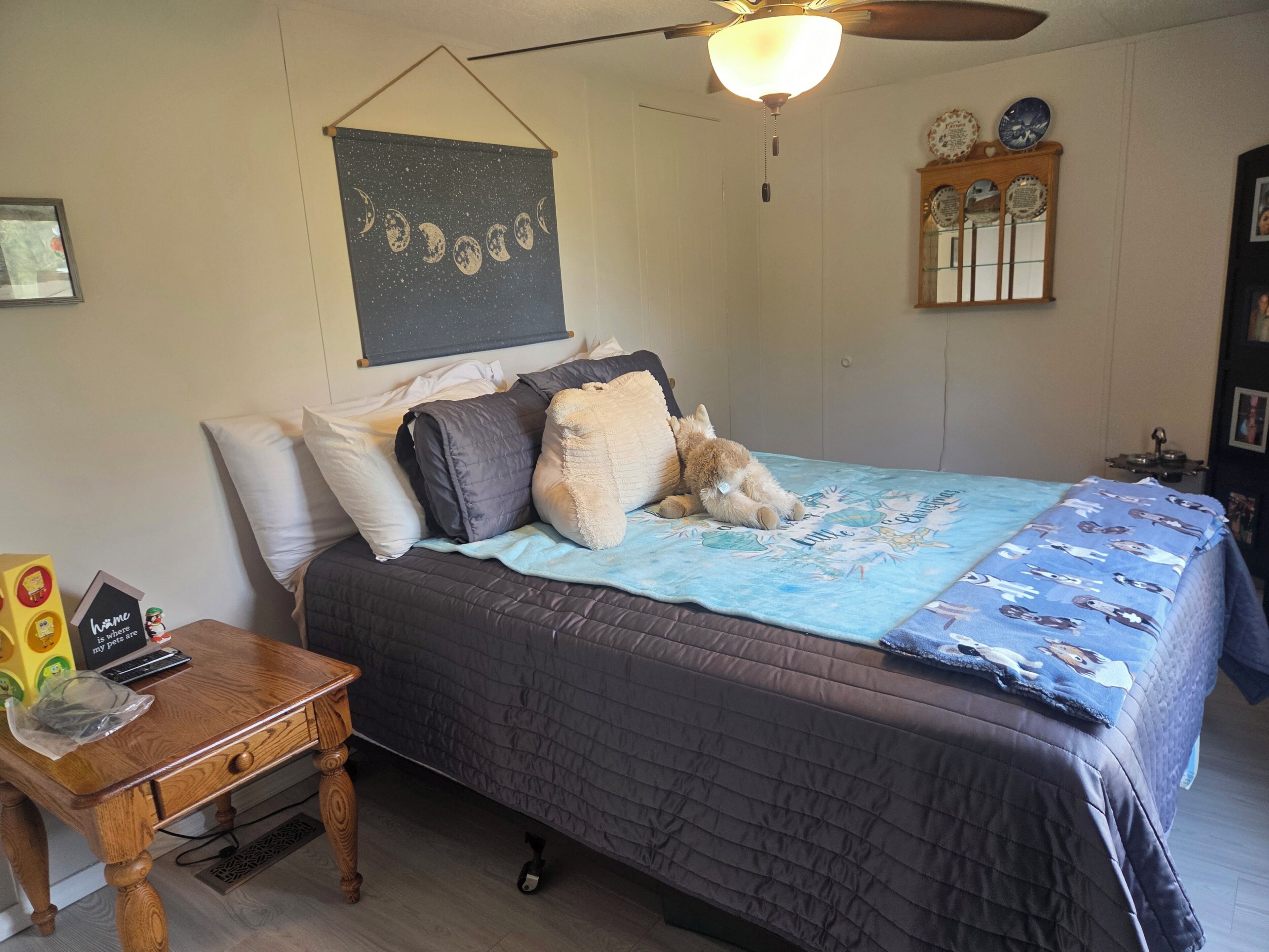 Een slaapkamer met een bed, kussens, een deken, een houten nachtkastje, een wandklok en een wandtapijt.